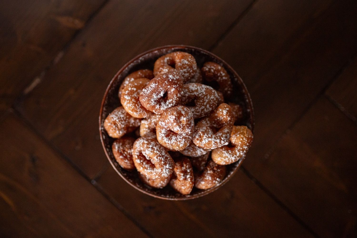 A bowl of donuts with powdered sugar on a wooden table.
