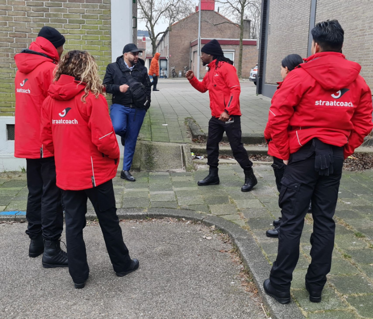Een groep mensen, sommigen in rode jassen, op een stoep, mogelijk in gesprek.