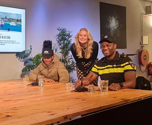 Drie mensen zitten lachend aan een tafel. Een vrouw en twee mannen, van wie één een politie-uniform draagt, voor een scherm en een potplant.