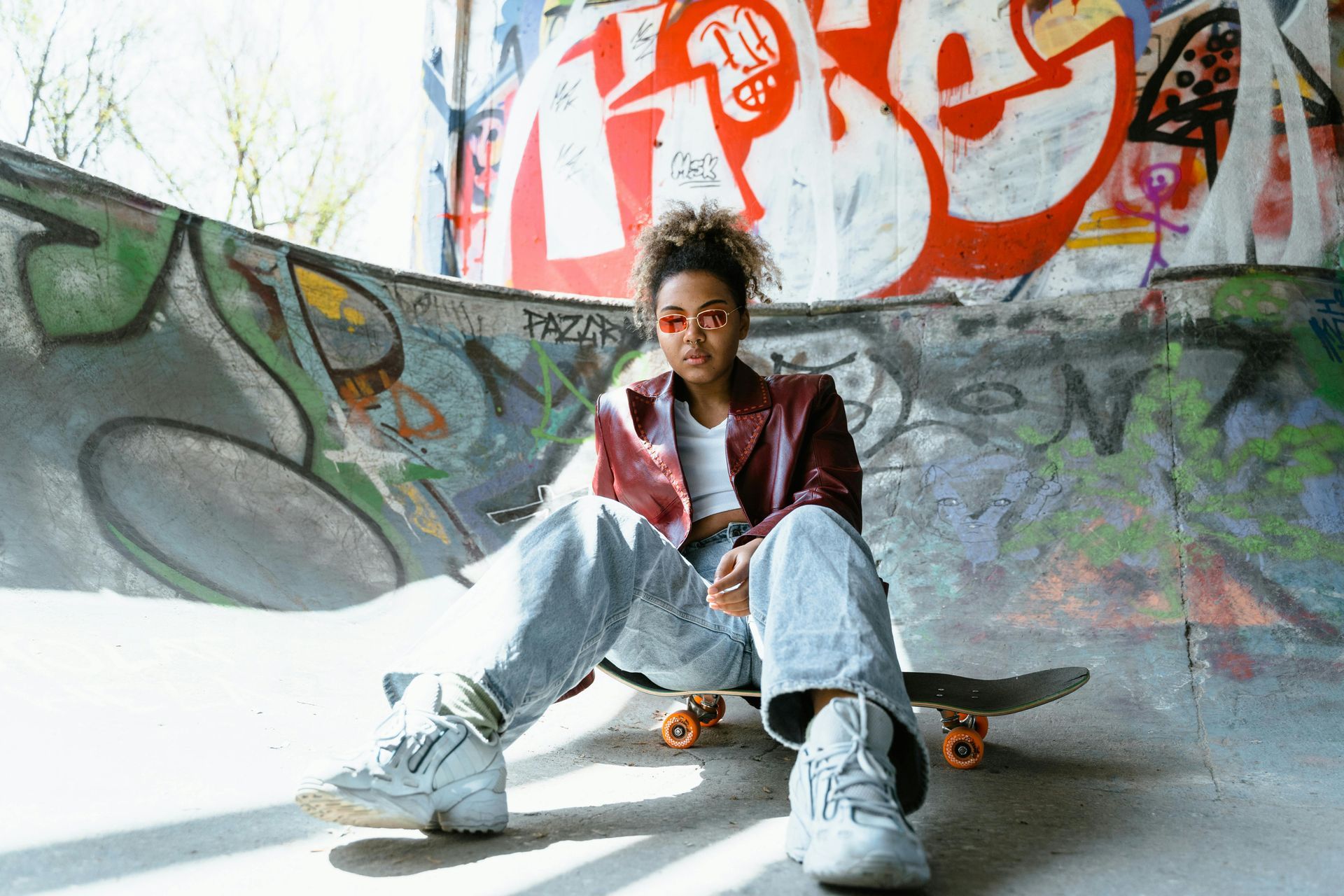 Vrouw zit op een skateboard in een skatepark, gekleed in een bordeauxrode jas en een oversized spijkerbroek, omringd door graffiti.