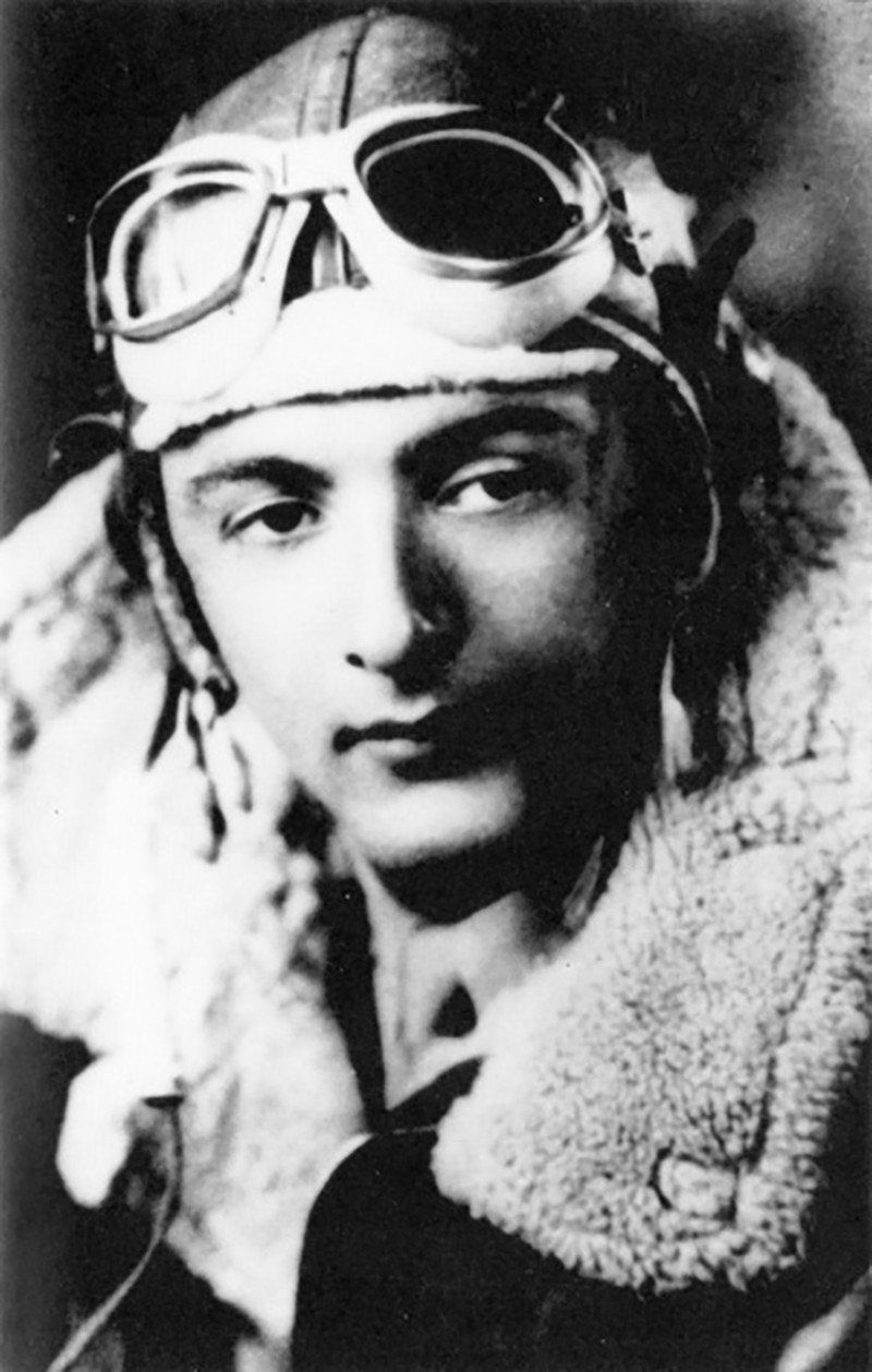 Young man in aviator gear, wearing goggles and a fur-lined jacket, looking at the camera.