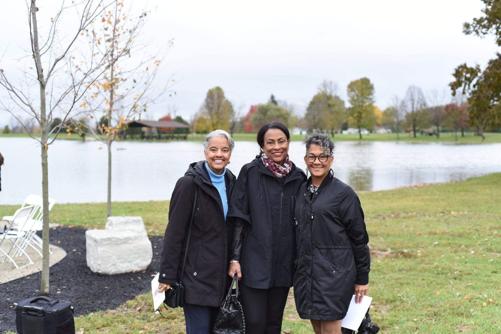Three women standing by a lake, wearing coats, smiling, with trees and a cloudy sky in the background.