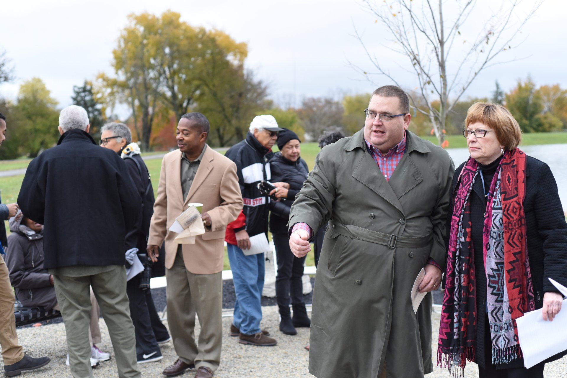 Group of people outdoors near a body of water. One man gestures while speaking, others listen. Cloudy sky.