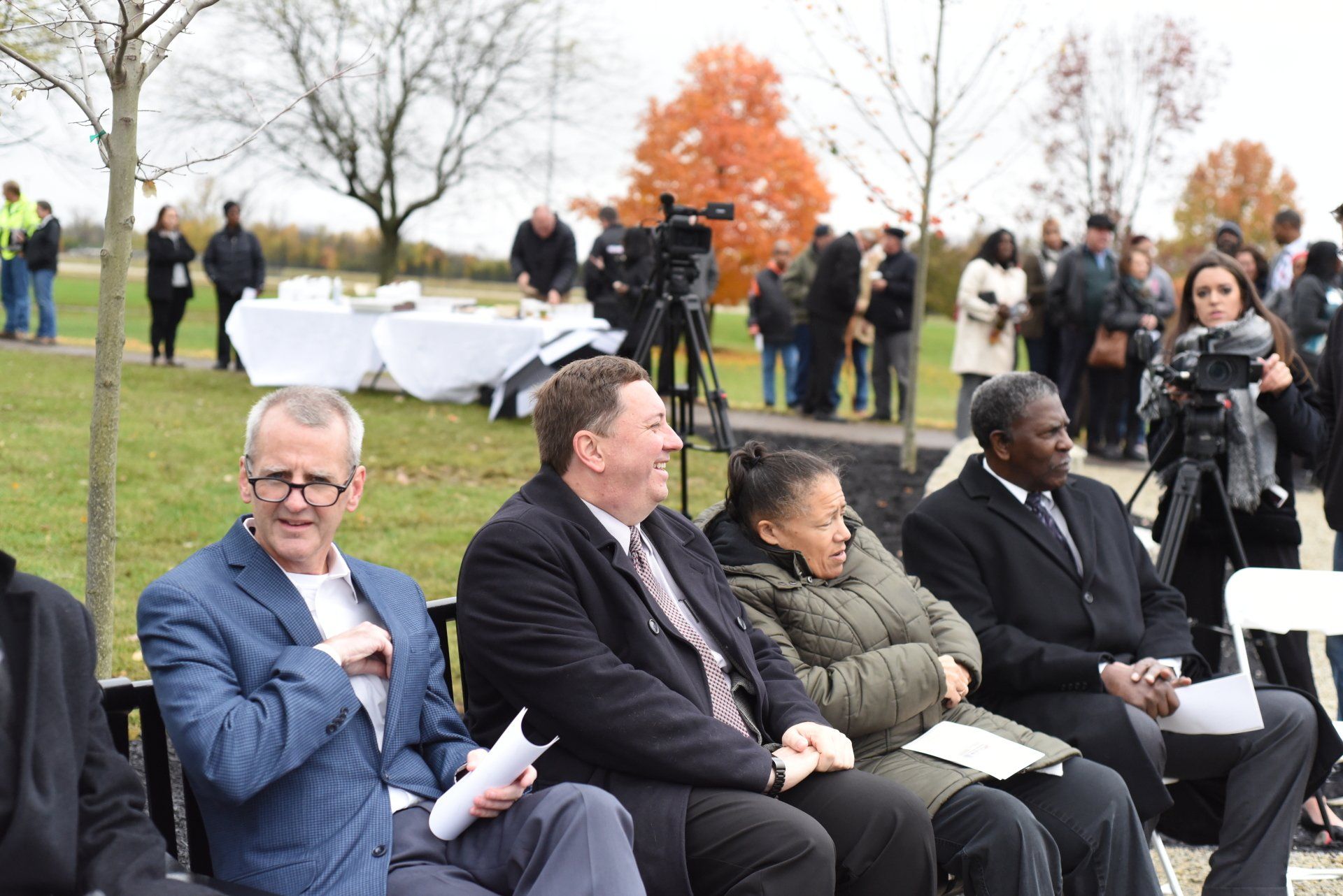 Group of people seated outdoors at an event, some smiling, with a backdrop of trees, lawn, and more attendees.