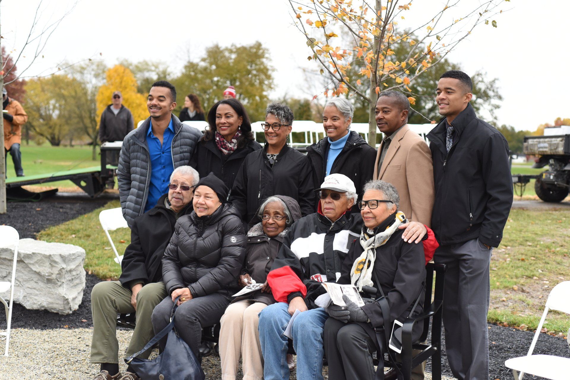 Group photo of Black people at an outdoor gathering. Some sit, others stand, smiling at the camera. Fall foliage in background.