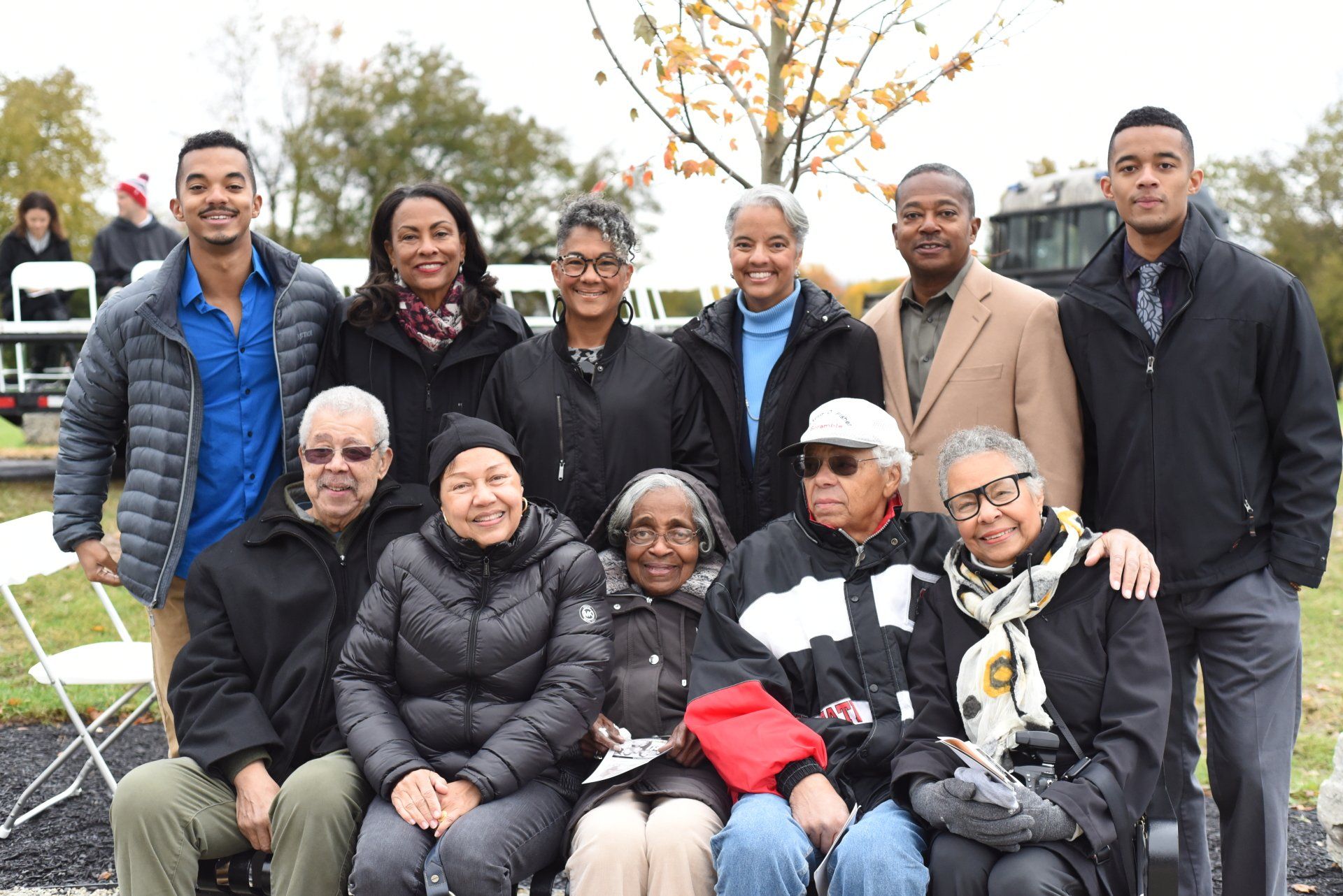 Group of diverse people smiling outdoors. Some are seated, others standing; autumn setting.
