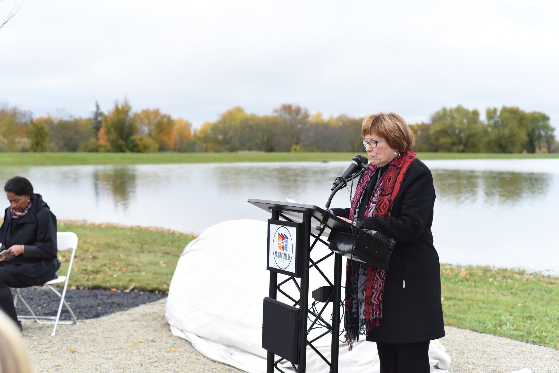 Woman speaking at a podium near a body of water on a cloudy day.