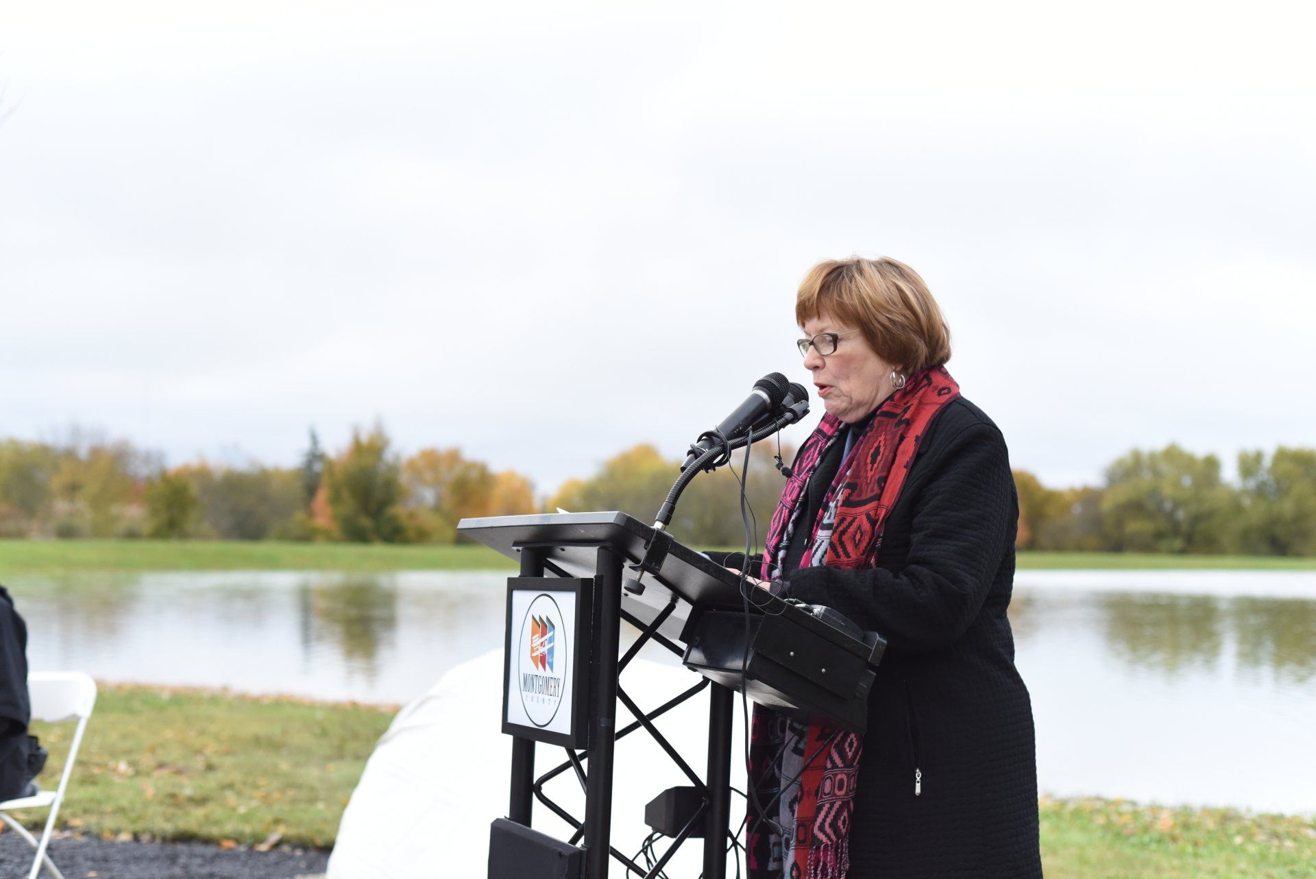 Woman speaking at a podium outdoors near a body of water. Fall foliage in background.