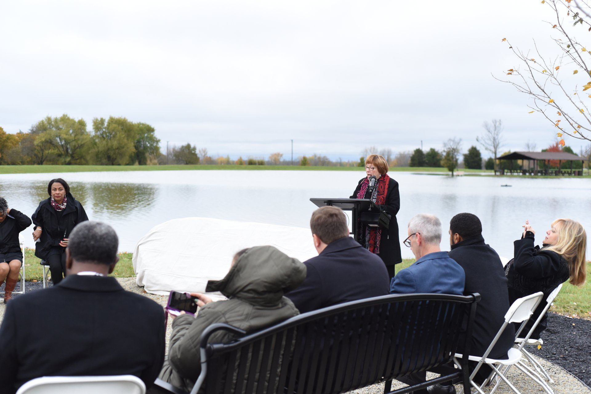 A woman speaks at a podium by a lake; an audience sits nearby.