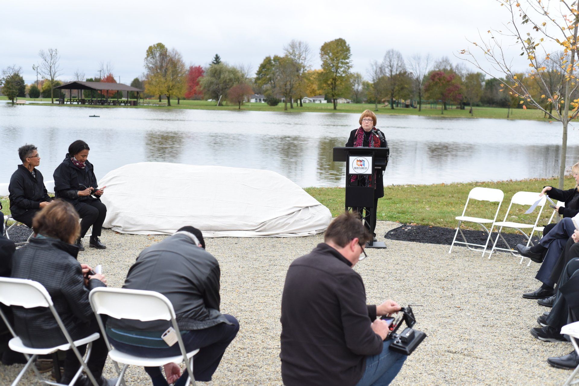 Woman speaking at podium, surrounded by people, lake in background, overcast day.