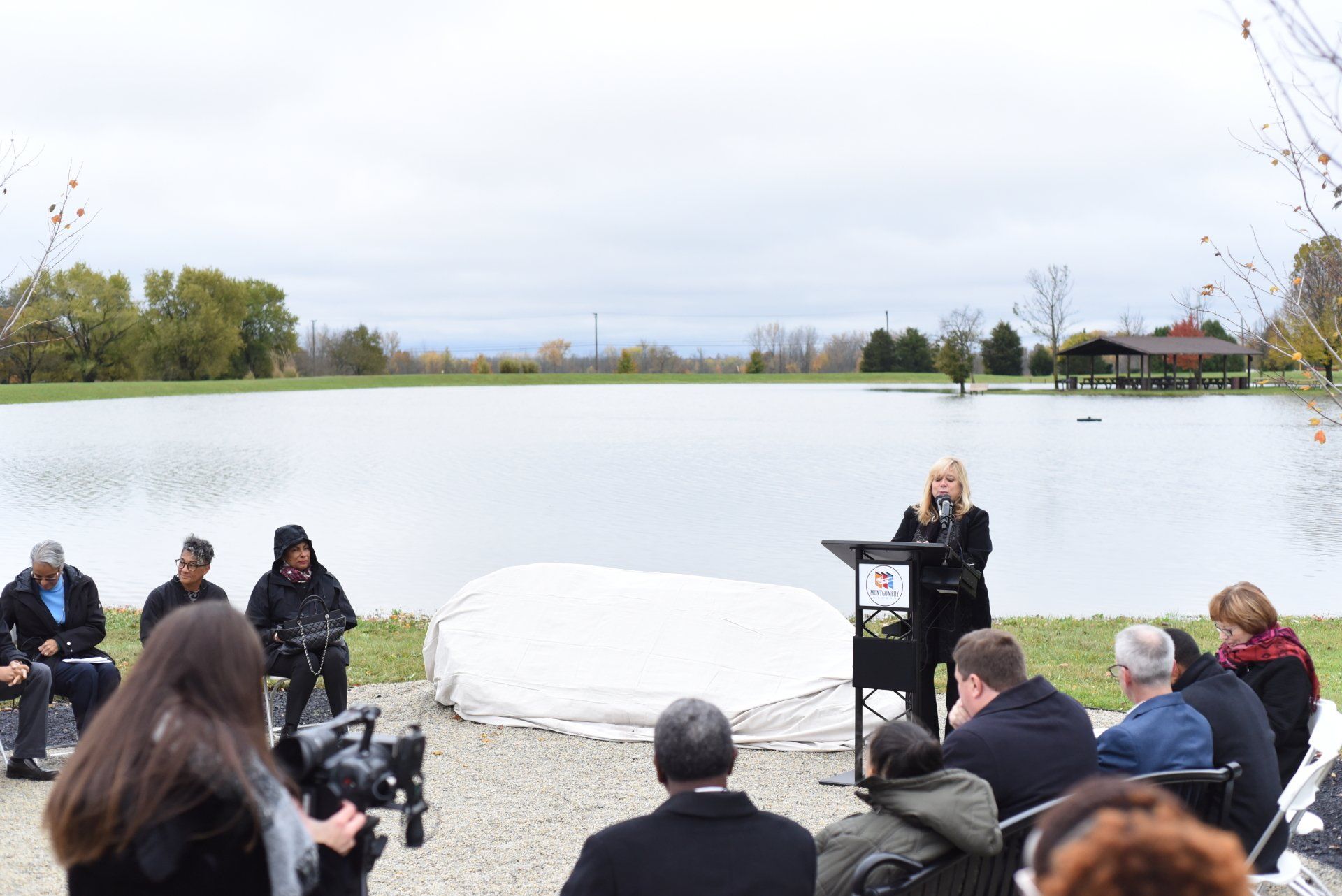 Woman speaking at podium near a lake, surrounded by seated people outdoors under an overcast sky.