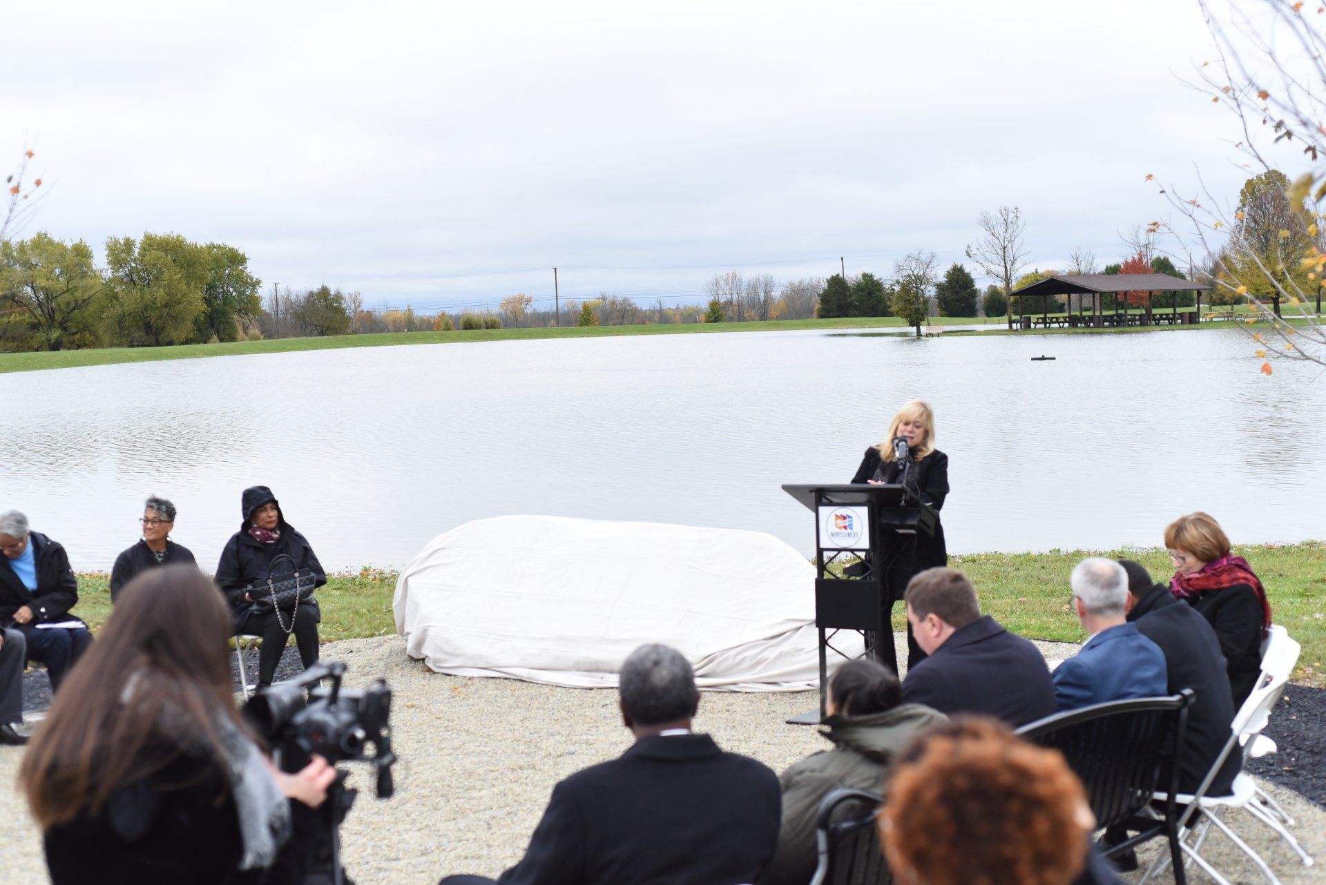 Woman speaking at a podium near a lake, surrounded by people. A covered object is on the ground. Cloudy day.