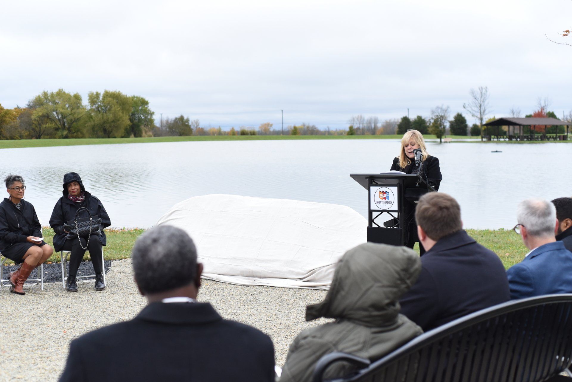 Woman speaking at podium; people seated outdoors by a lake.