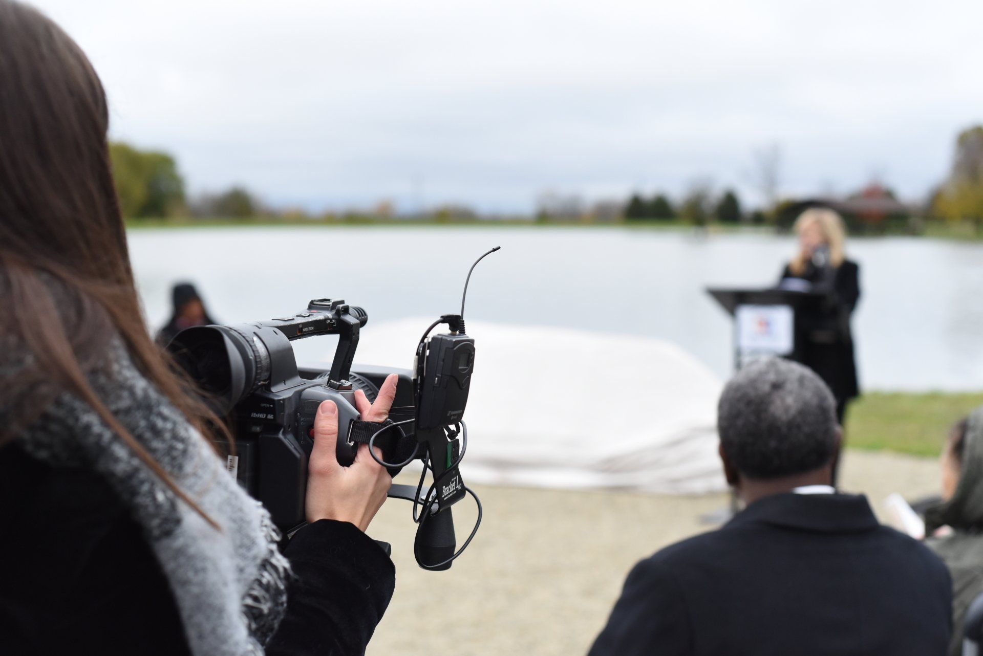 Person films a woman speaking at a podium by a lake; other people attend.