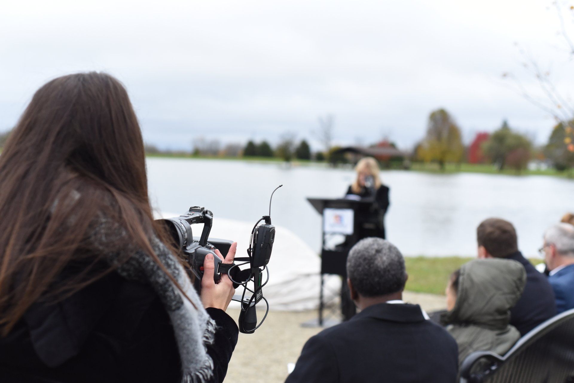 A cameraperson films a woman speaking at an outdoor podium. People listen on a cloudy day near water.