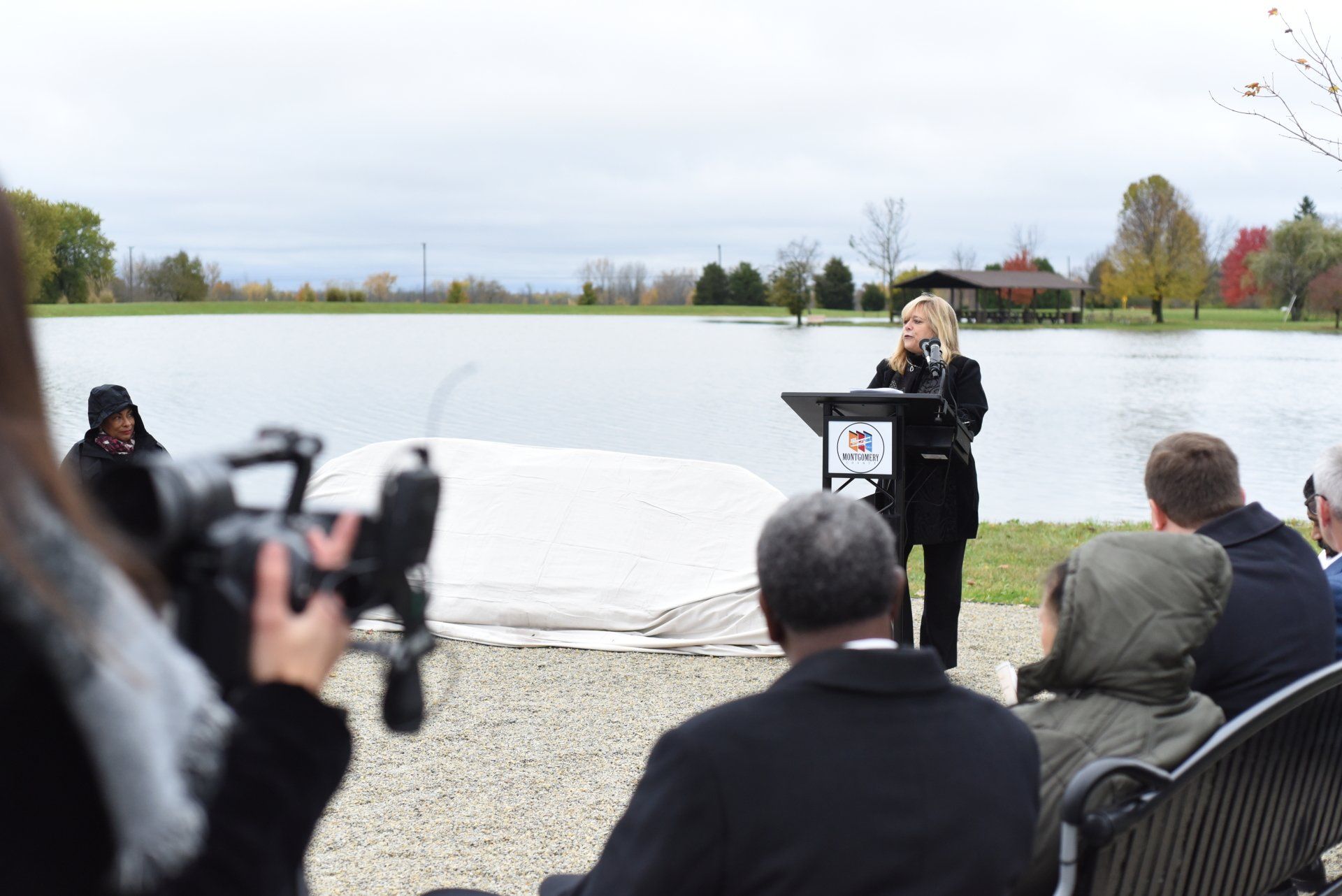 Woman speaking at podium near a lake, flanked by a small audience, trees, and cloudy sky.