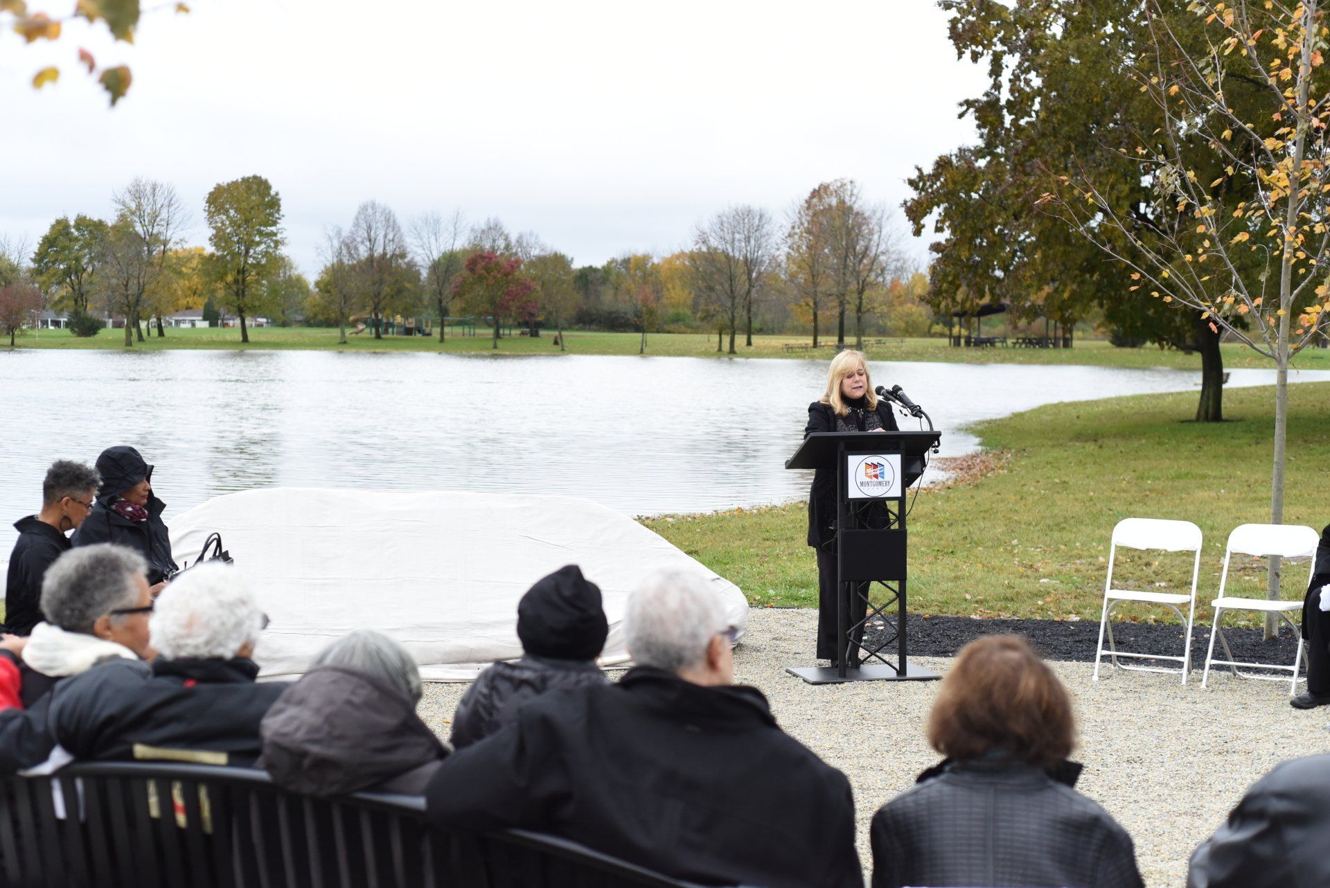 Woman speaking at podium, lakeside. Audience seated on benches, trees and water in the background.