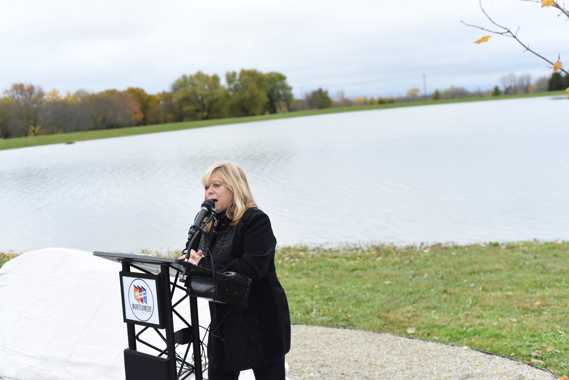 Woman speaking at a podium near a lake, wearing a black coat. Cloudy day.