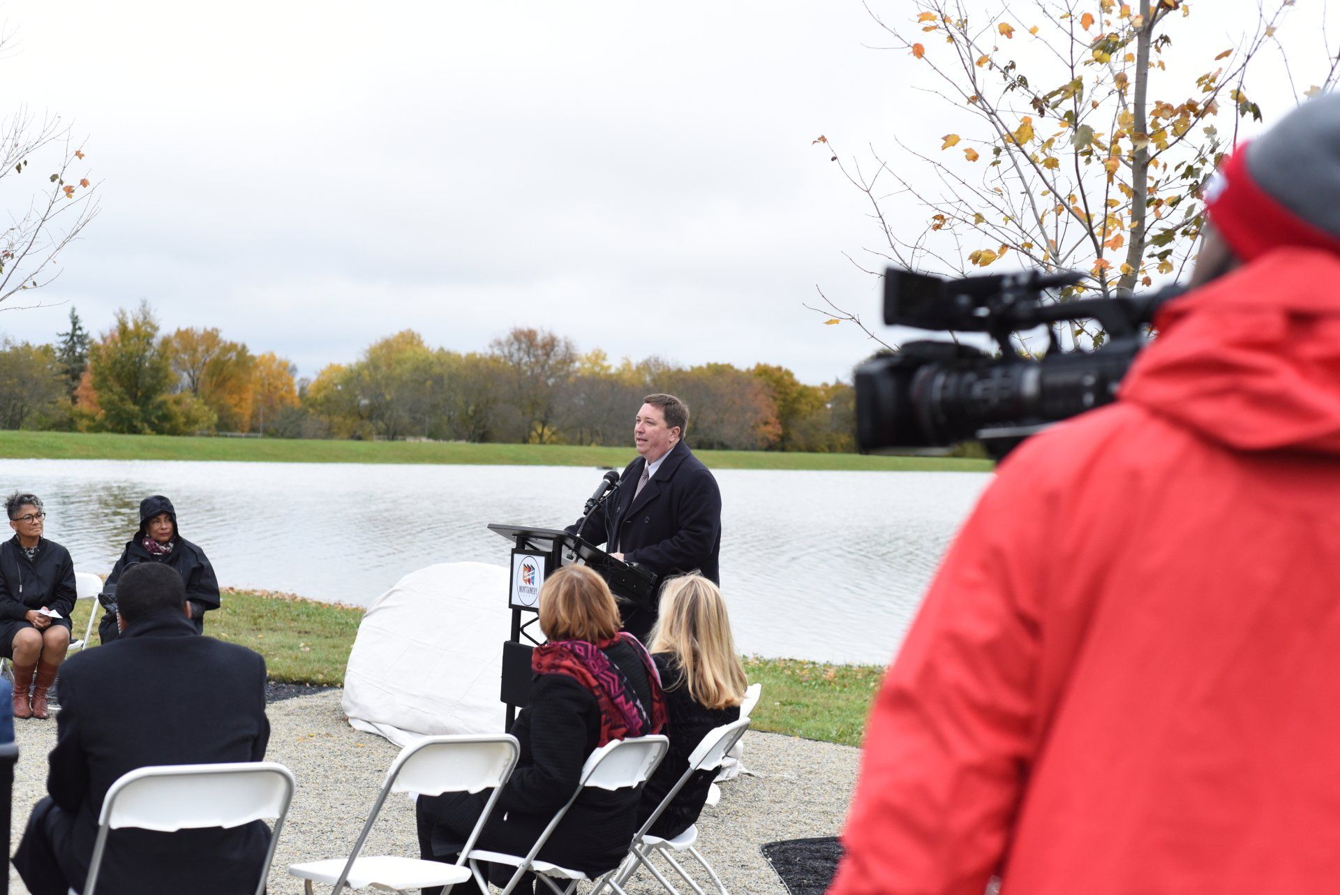 A person speaks at a podium outdoors by a pond, facing an audience and a camera operator.