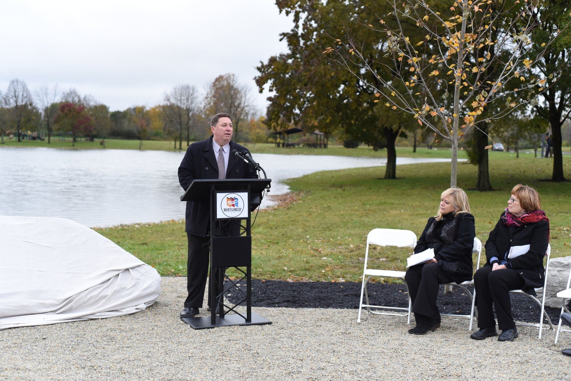 Man speaking at a podium near a lake, two women seated listening, fall day.