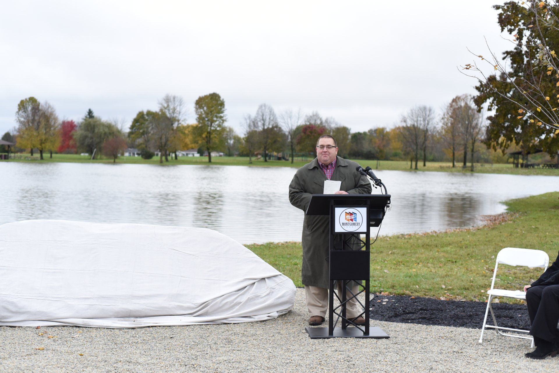 Man speaking at a podium outdoors, near water and covered object. Overcast sky, fall foliage in the background.