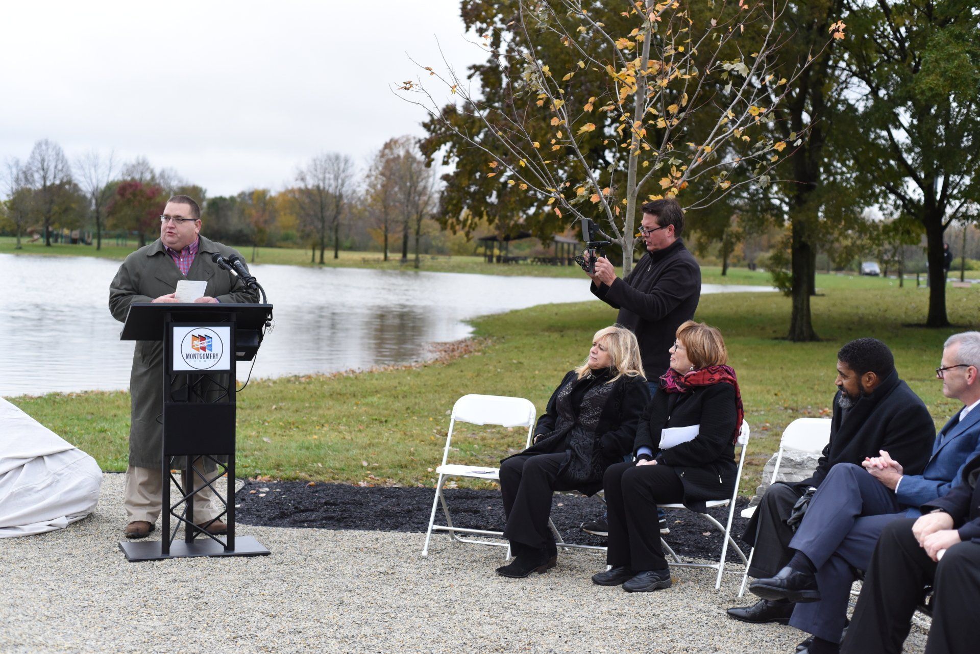 Man speaking at a podium near a lake, people seated, a man filming with a camera, trees in the background.