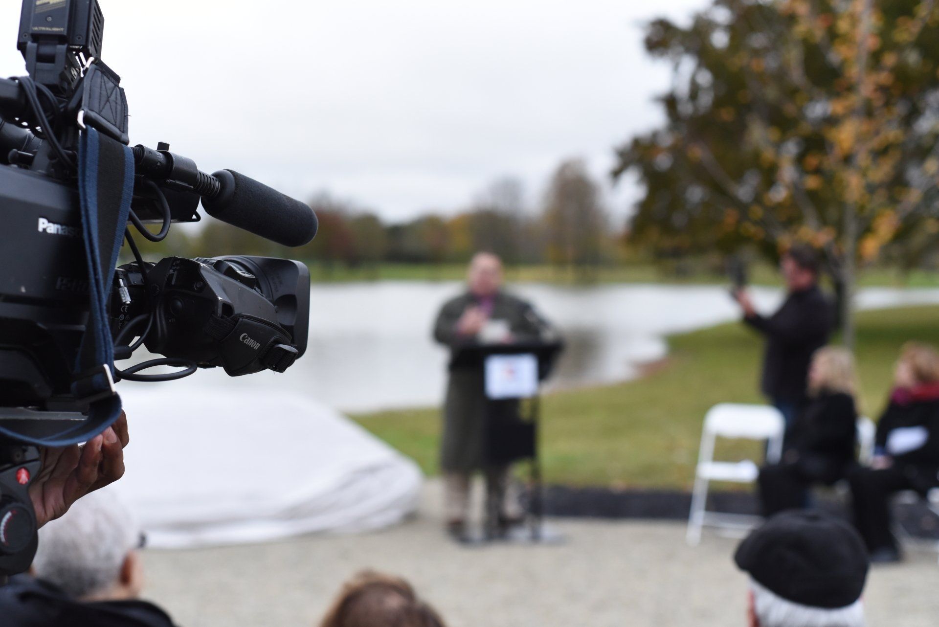 Camera filming a man speaking at a podium near a lake; several people watch in background.