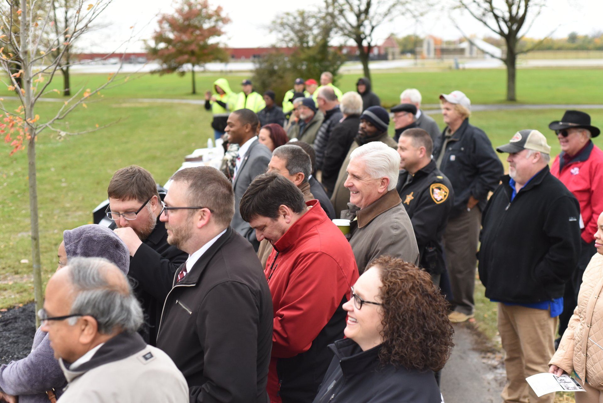 Group of people observing, likely at an outdoor ceremony. Many are looking at a central point, likely a planting. Cloudy day.