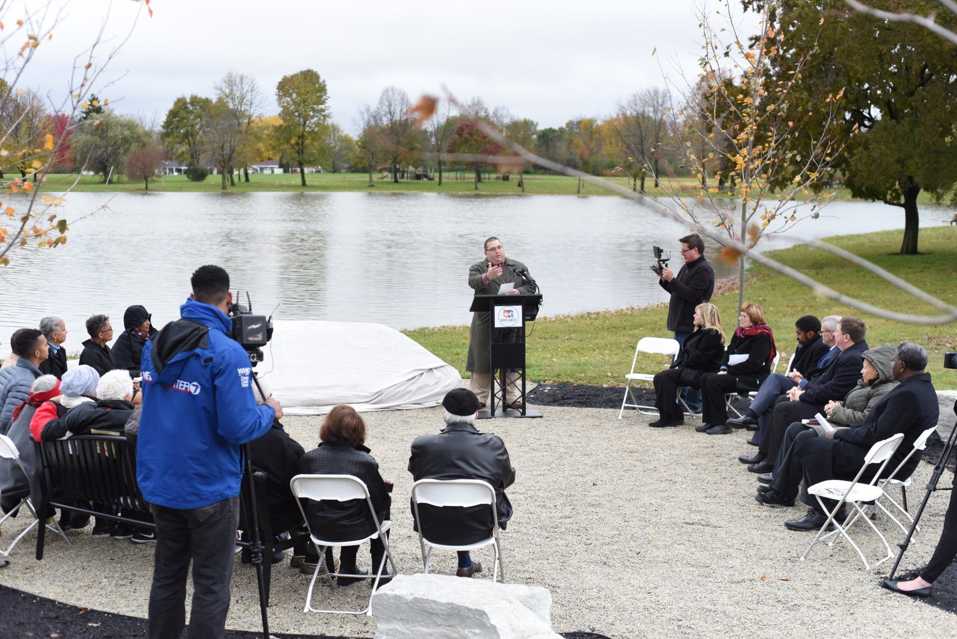 A man speaks at a podium outdoors, in front of a lake. A group of people listen; a reporter films.