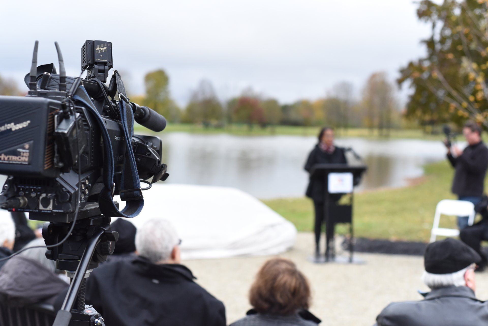 A person speaking at a podium near a lake, filmed by a camera on a tripod, with an audience.