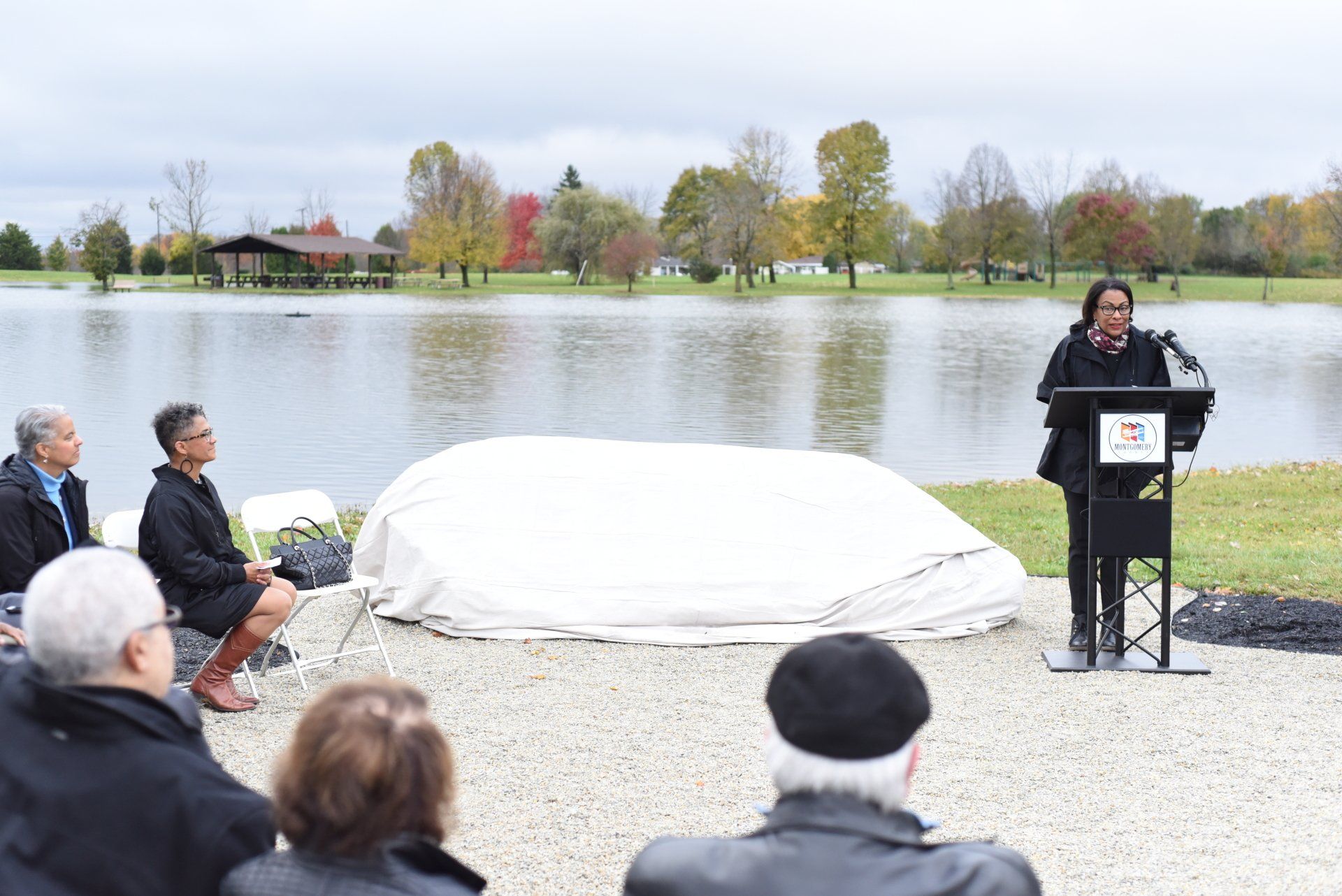 A woman speaks at a podium near a lake, unveiling an object covered in white. Other people listen.