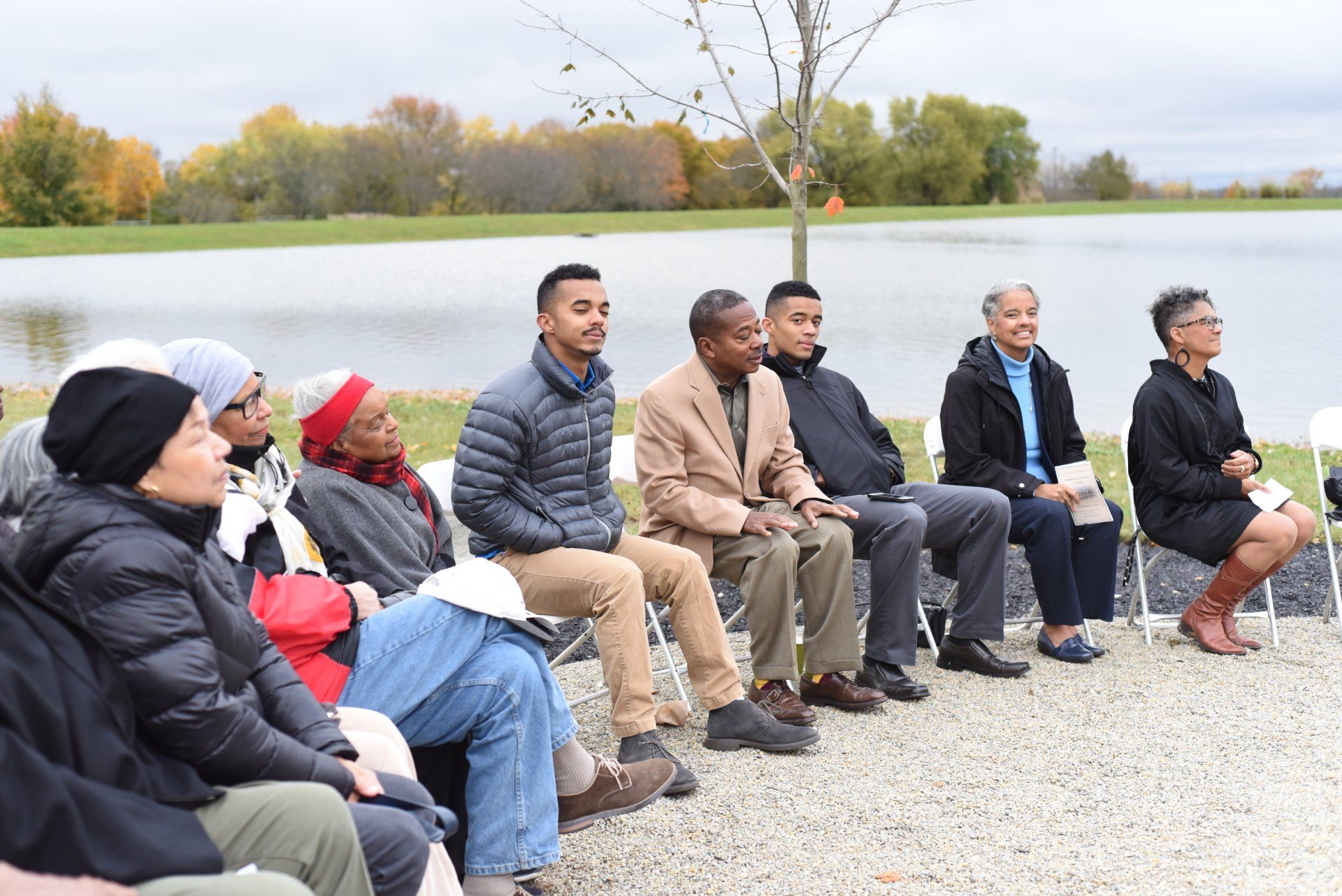 Group of people seated outdoors near a lake, some with eyes closed, likely a ceremony or event.