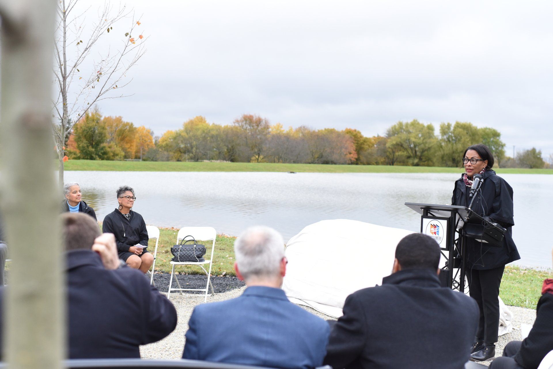 Woman speaking at a podium near a lake, addressing a seated audience on a cloudy day.