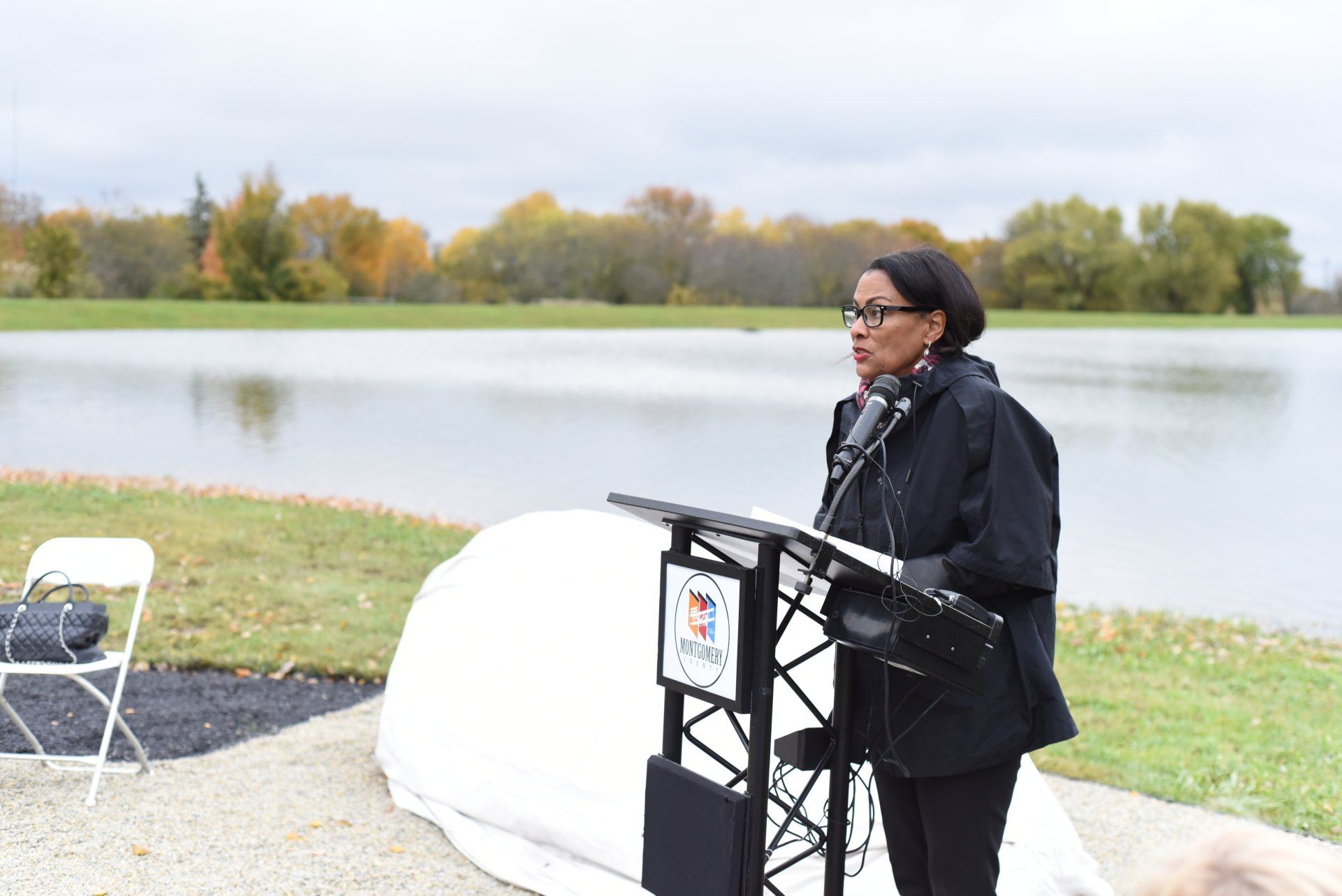 Woman speaking at a podium by a lake. Outdoors on an overcast day.