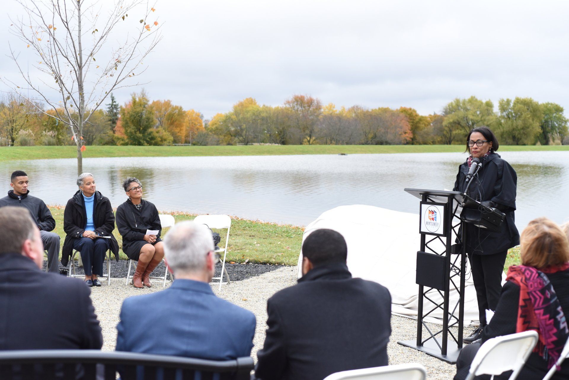 Woman speaking at podium by a lake, several people seated nearby, fall foliage in the background.