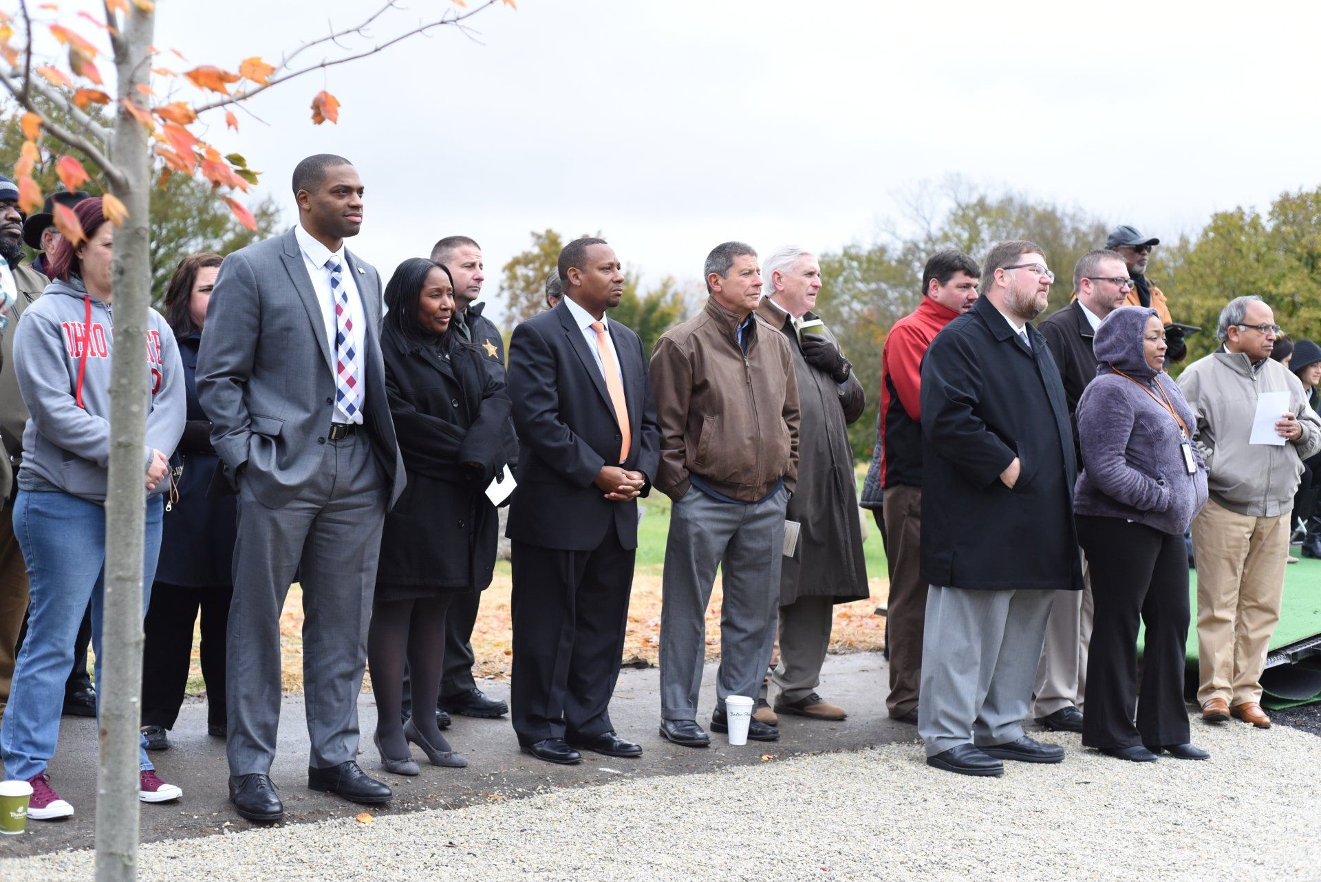 Group of people, diverse in age and dress, standing outdoors, watching.