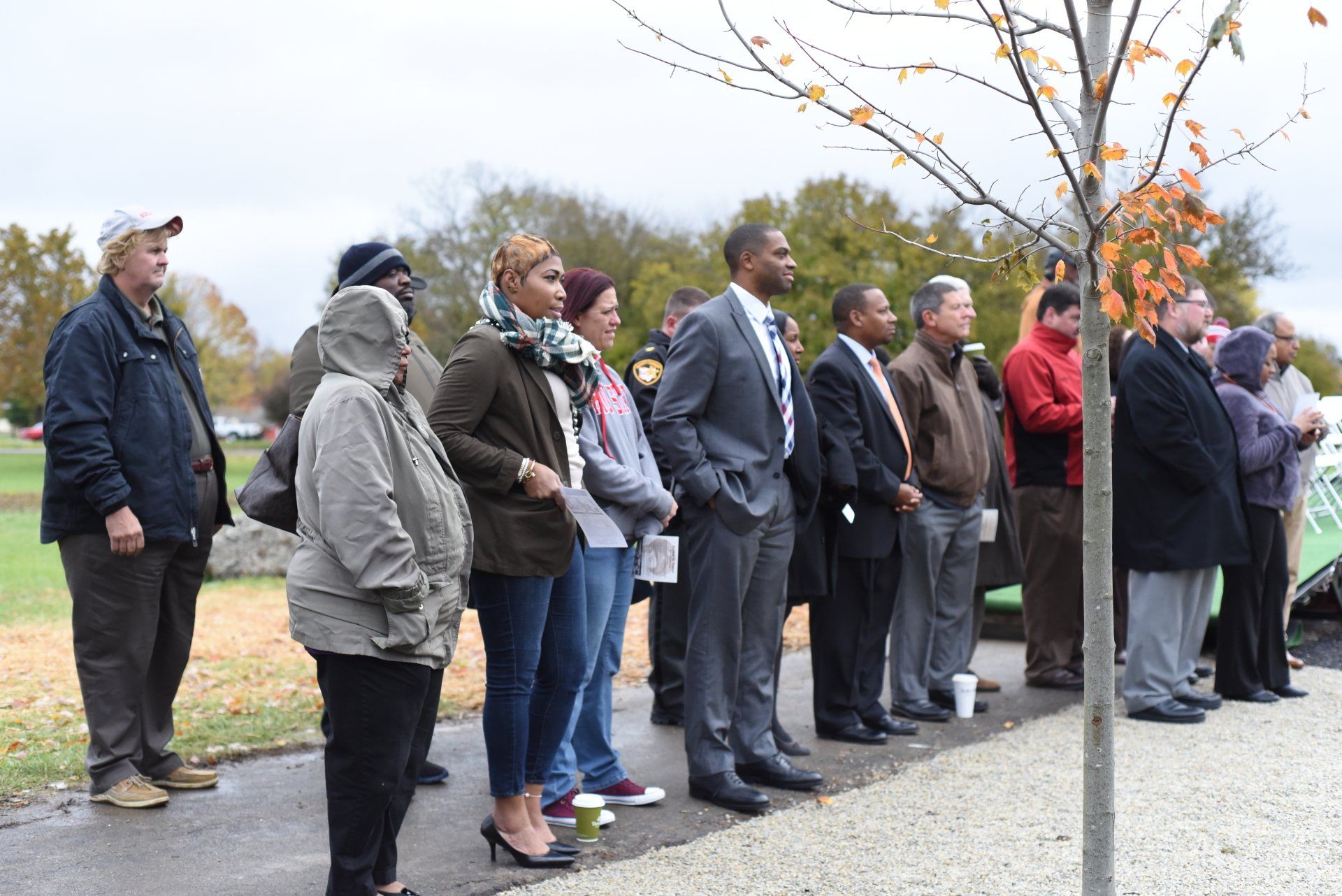 Group of people standing outside, looking towards something; overcast day.