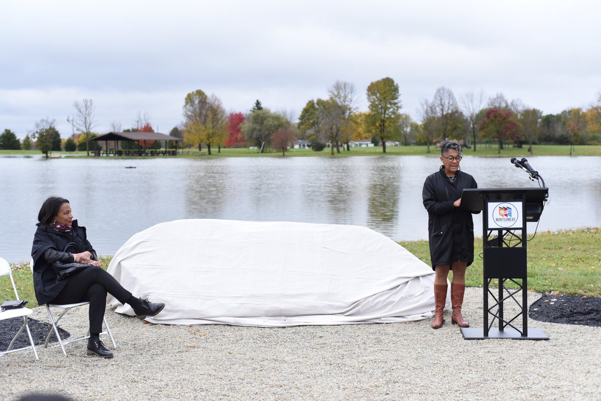 Two women at an outdoor event near a lake; one speaking at a podium, the other seated.