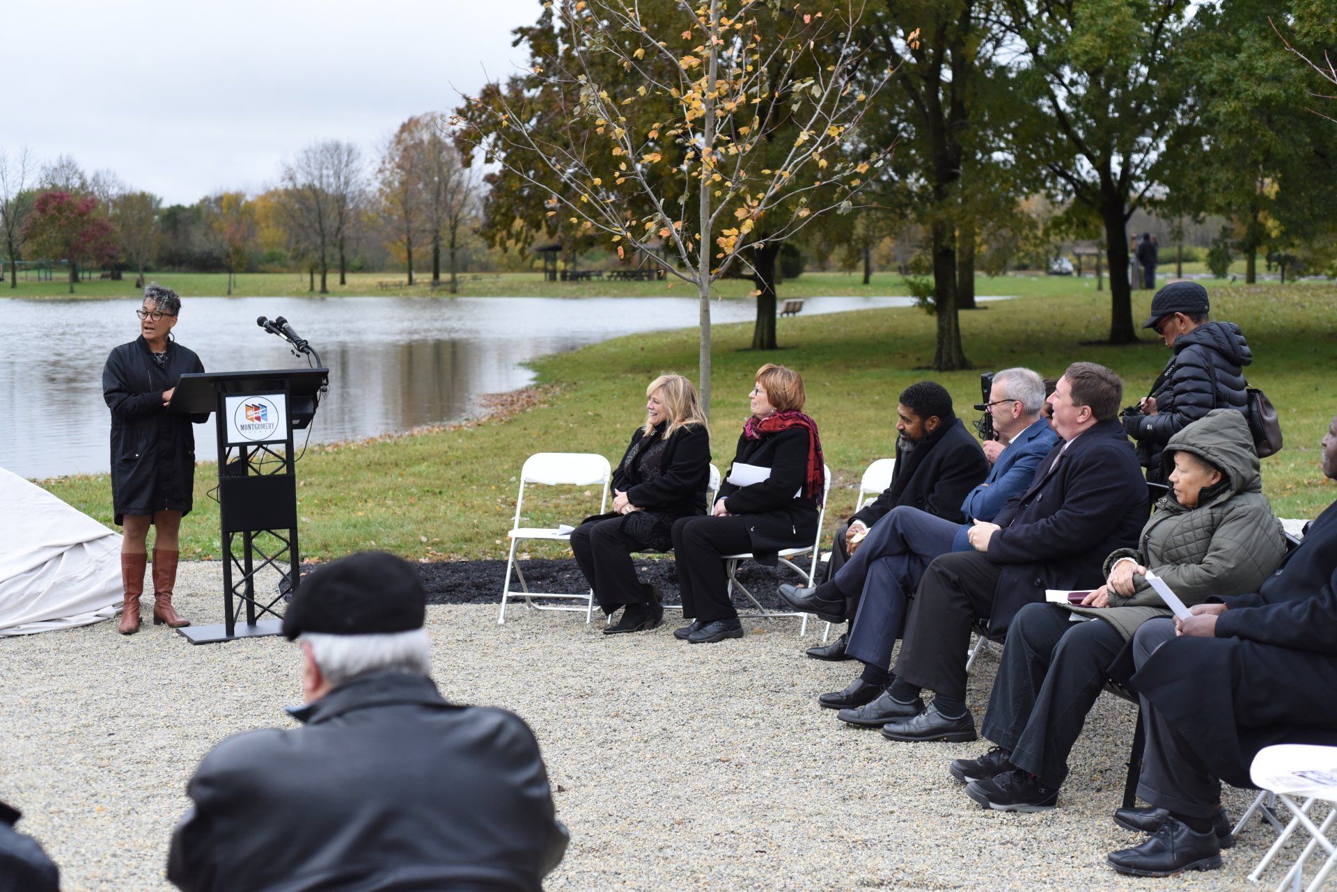 Woman speaking at outdoor event, with audience seated by a lake; overcast sky, fall foliage.