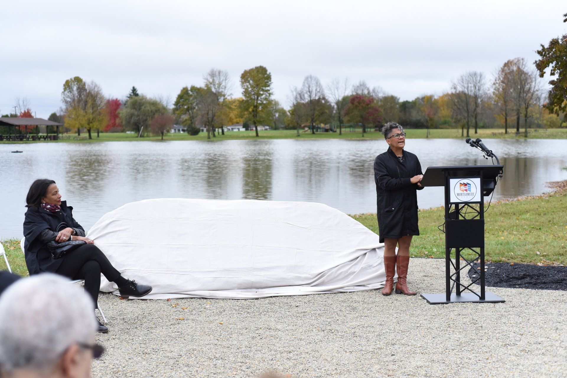 Woman speaking at a podium by a lake. Another woman sits to the side. Overcast day.