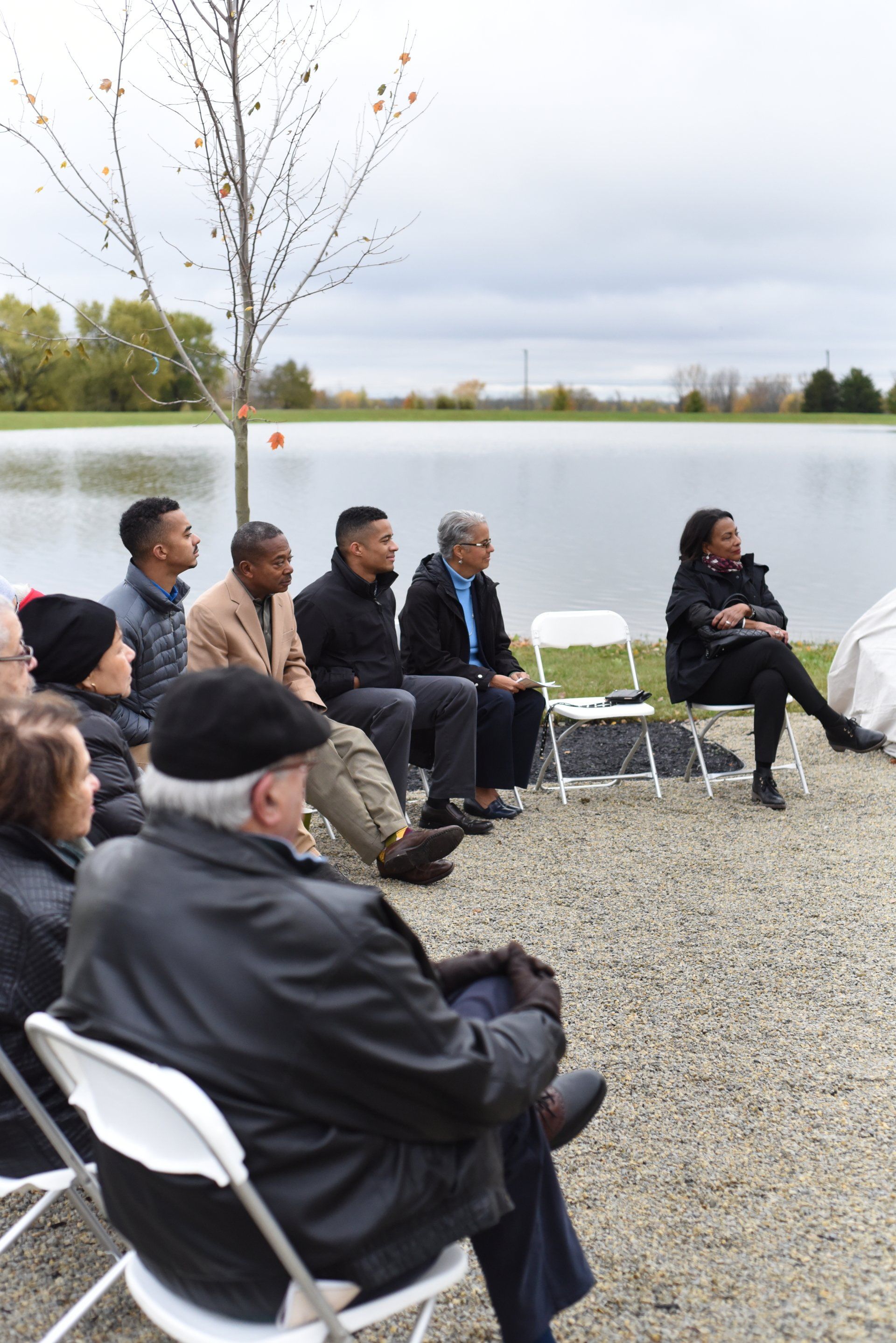 Group seated outdoors by a lake, listening to a woman speaking; overcast sky.