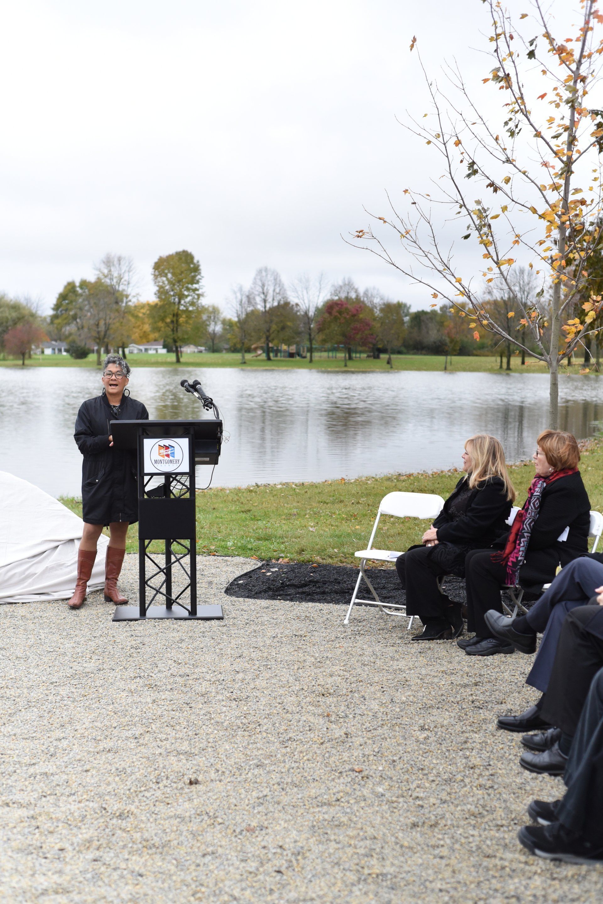 Woman speaking at a podium by a lake, with audience members seated. Overcast day.