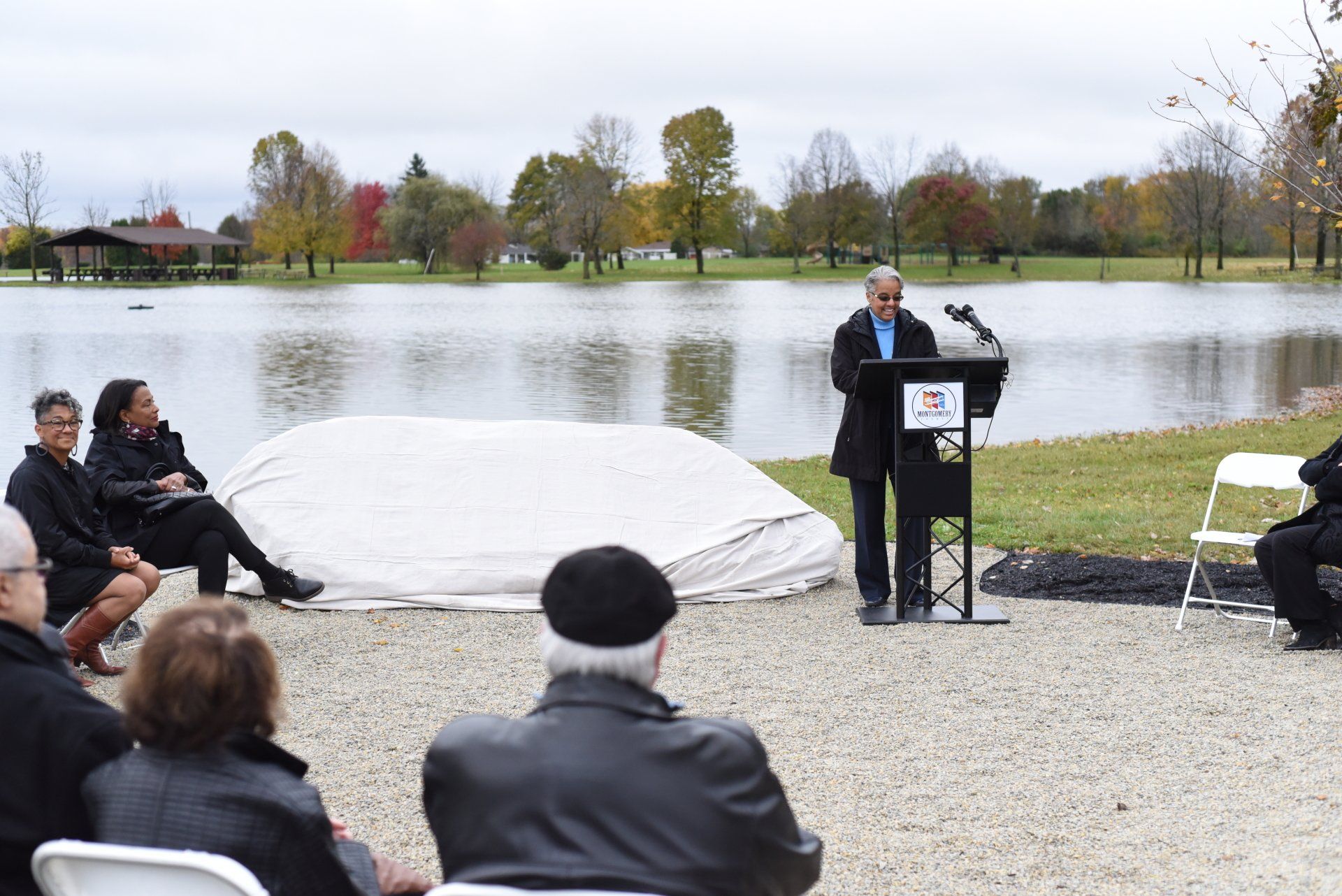 Man speaking at a podium by a lake; audience seated, fall foliage in the background.