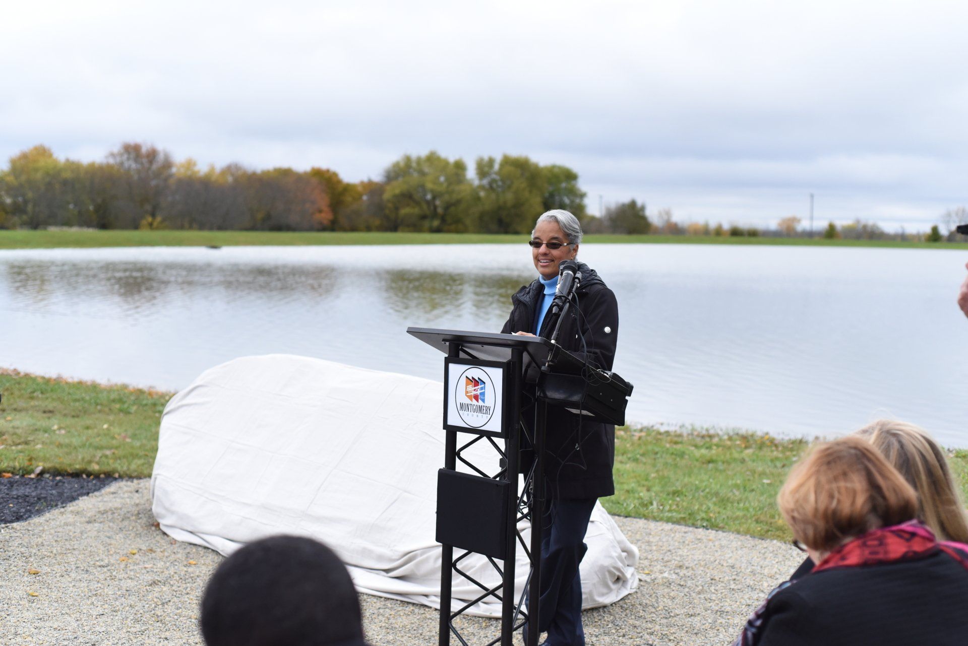 Man speaking at podium, covered object in front of lake; group of people in attendance.