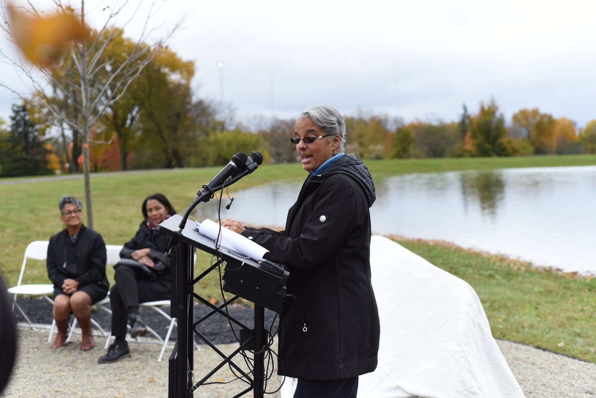 Woman speaking at podium outdoors, two women seated behind her near a lake.