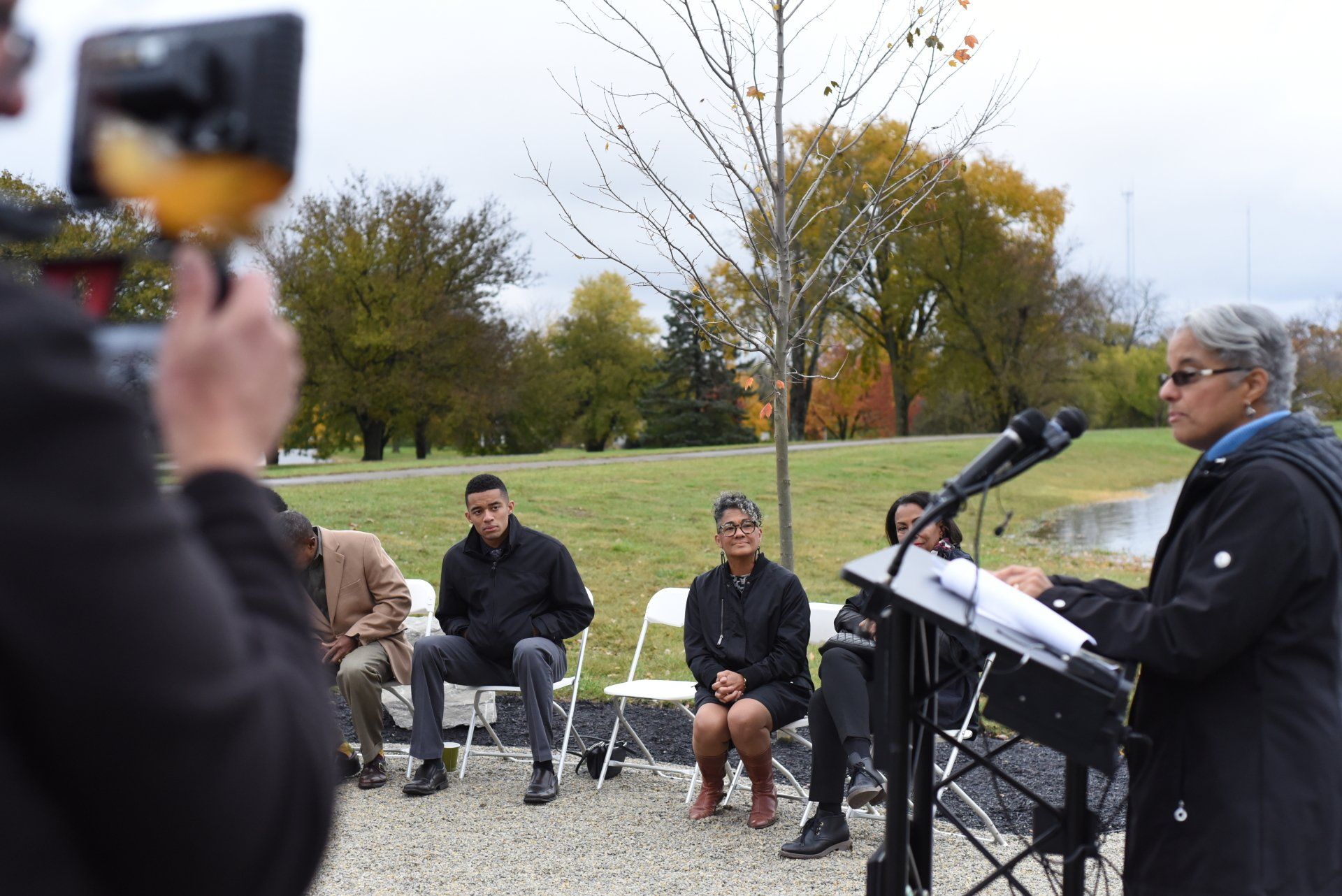 Woman speaks at a podium outdoors. People seated in front of a park. A person records with a camera.