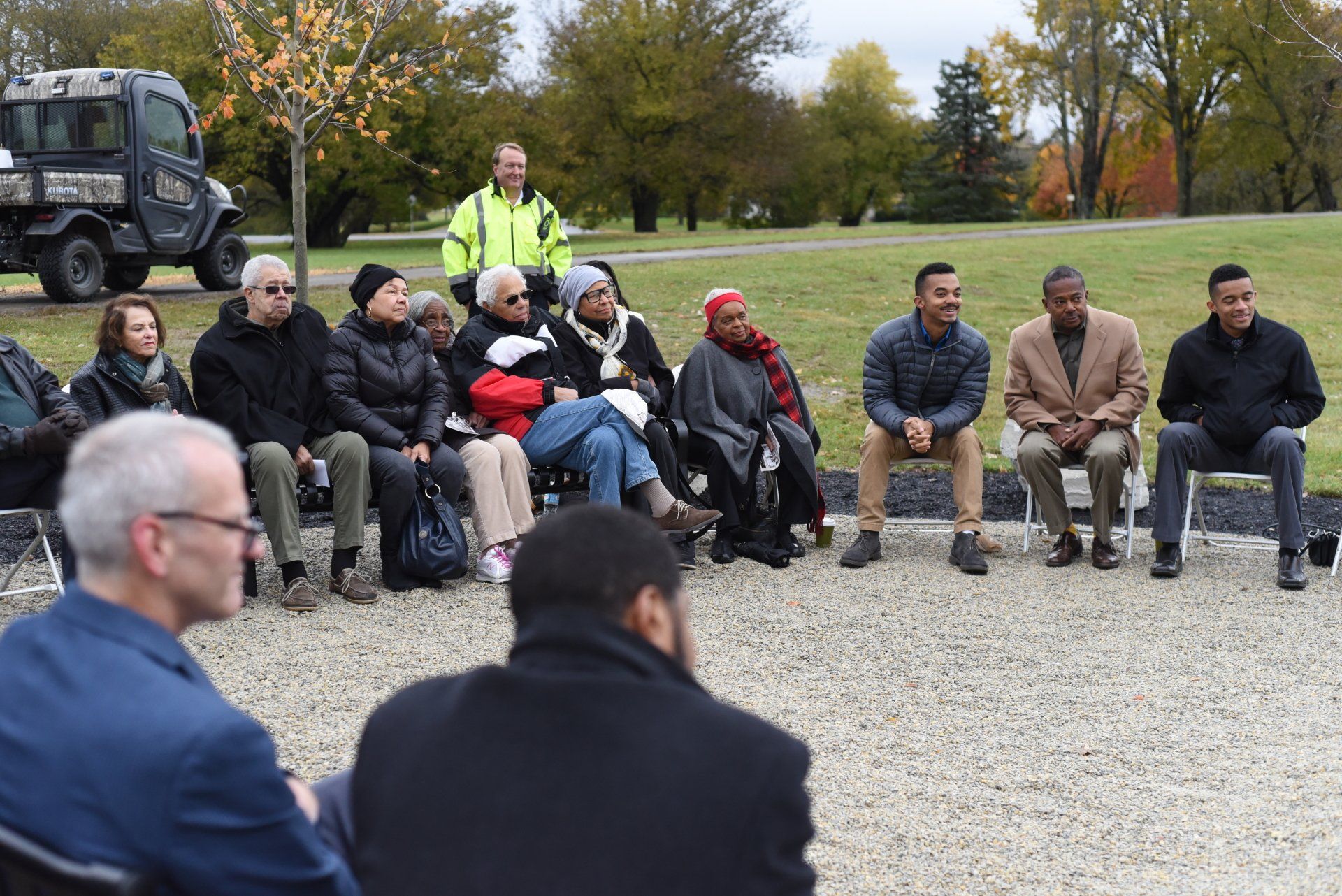 Group of people sitting outdoors, some looking at others; one man in a neon vest stands nearby.