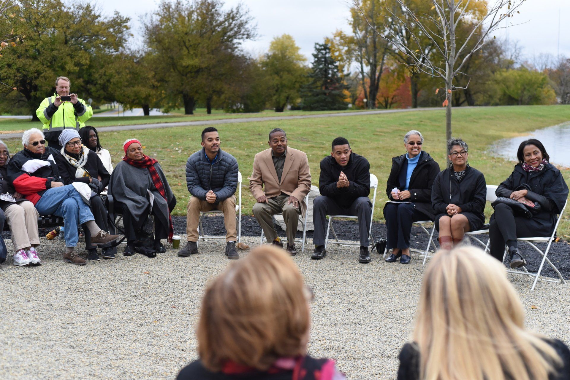 A group of people sits in a circle outdoors. Some are speaking. Fall foliage and pond in the background.
