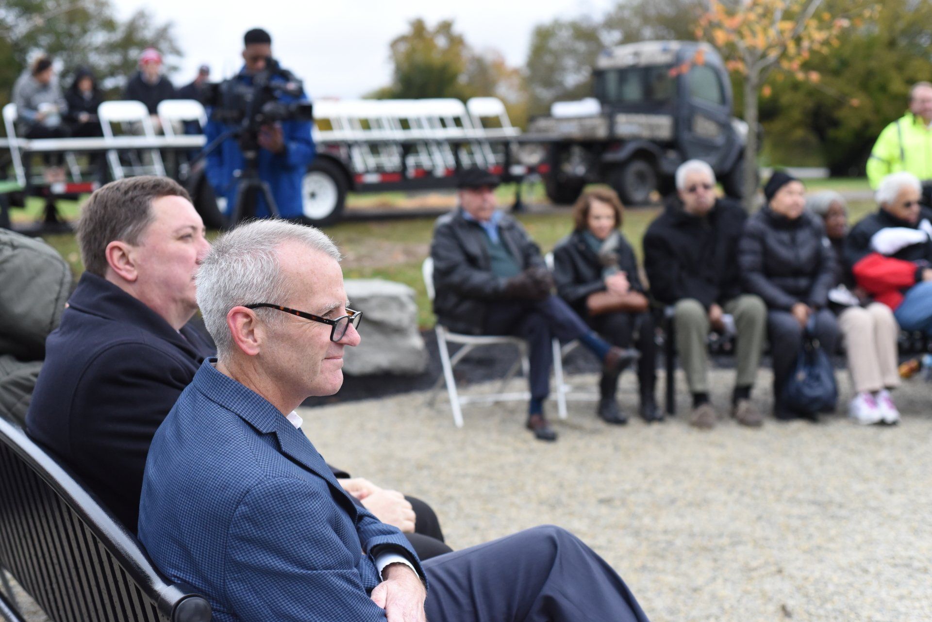 Men and seated group at an outdoor event. Man in blue suit smiles, others observe.