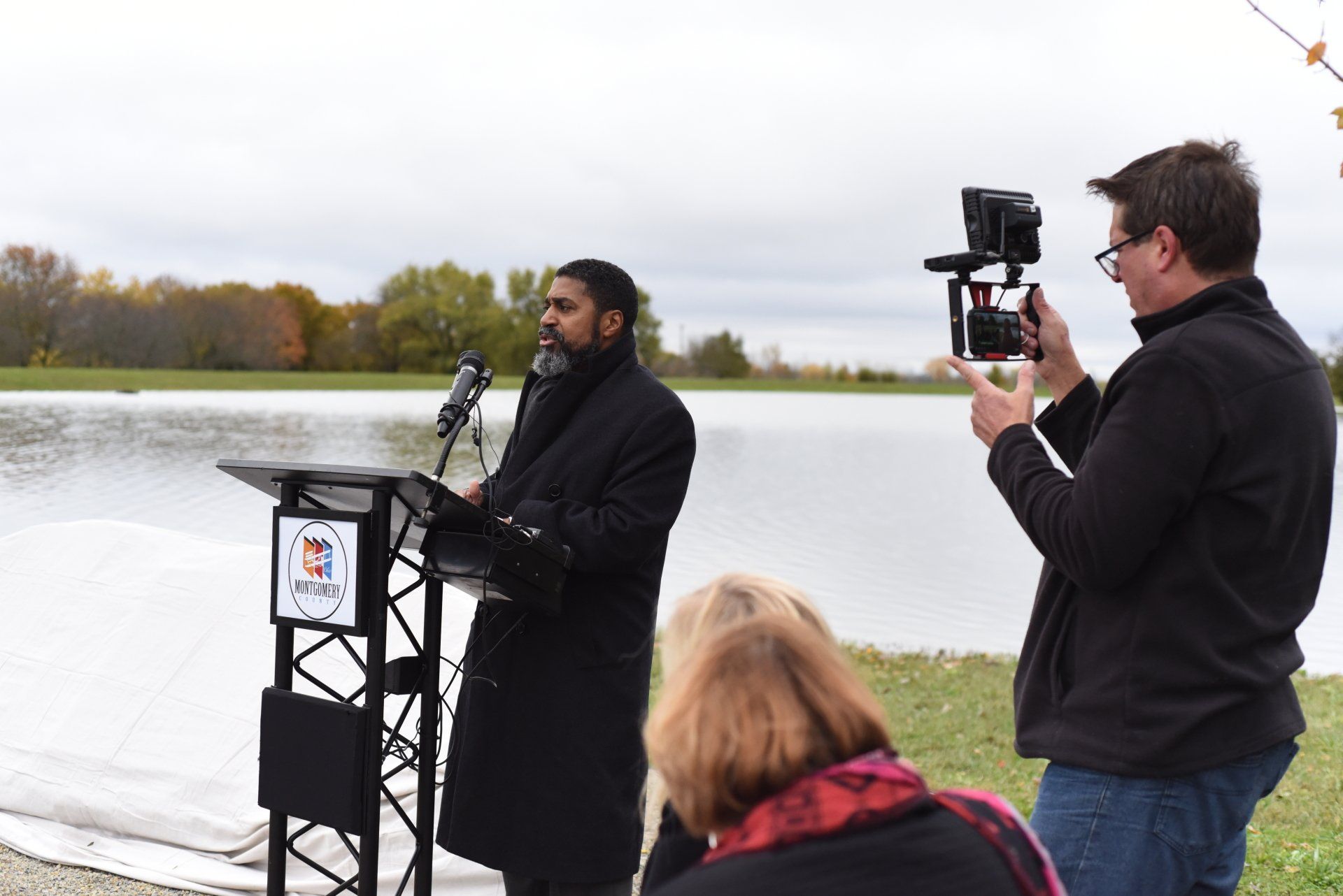 Man in black coat speaks at a podium outdoors, lake in background, filmed by a man with a camera.